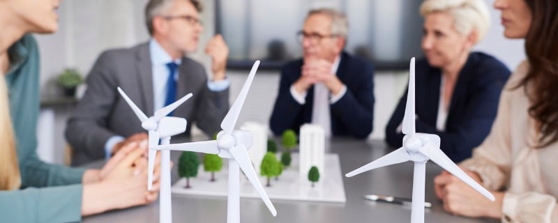 Trois modèles réduits d'éoliennes posés sur une table avec une réunion de travail qui se tient dans le fond de l'image