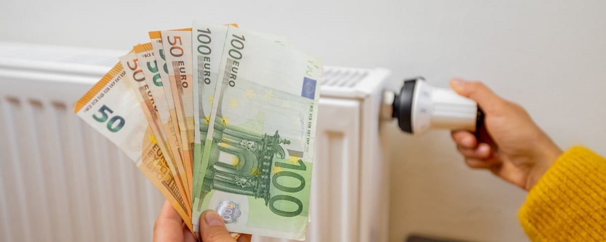 Main d’une personne tenant des billets de banque en euros près d'un chauffage à la maison, concept de ressources énergétiques coûteuses et de la crise énergétique en Europe.