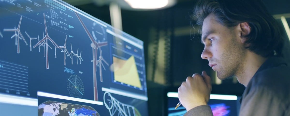 Un jeune homme aux cheveux longs, travaillant de nuit devant un grand écran d'ordinateur montrant les plans d'une éolienne (parc éolien).
